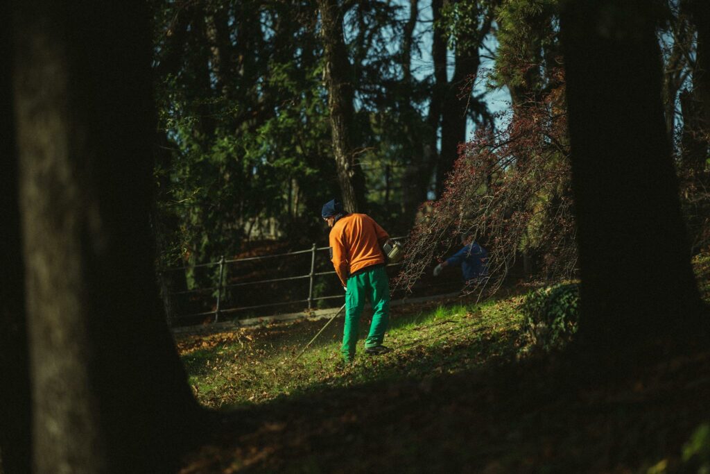 A man in vibrant clothing mows grass in a sunlit park surrounded by trees.